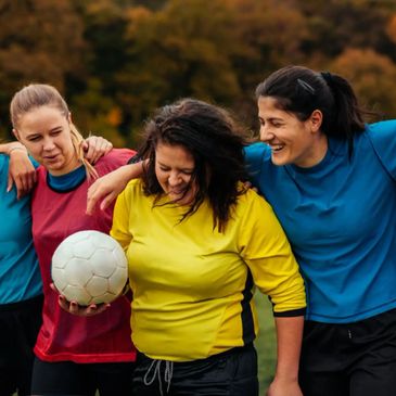 Three women in colorful sportswear smiling with a soccer ball outdoors.