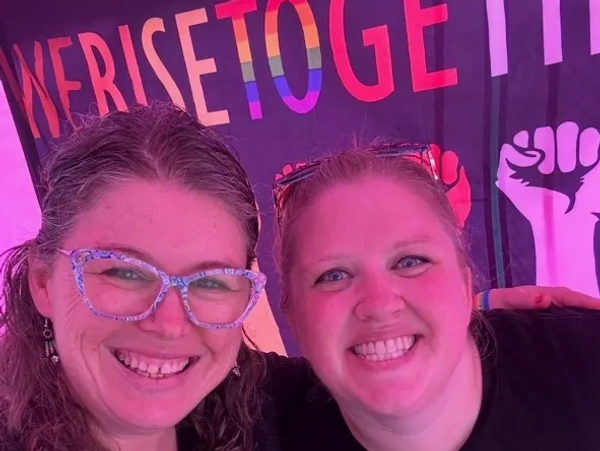 Two smiling women wearing matching scissor-printed shirts in front of a colorful banner.