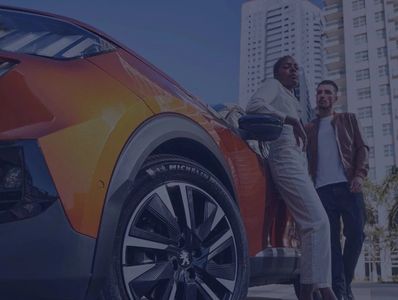 Two people standing next to an orange Peugeot car in an urban setting.