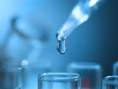 Close-up of a dropper releasing liquid into a test tube in a lab setting.