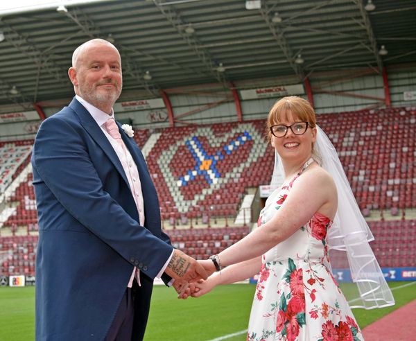 Bride amd groom holdings hands in front of Heart of Midlothian stadium 