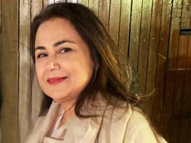 A smiling woman in a beige coat standing by a wooden wall.