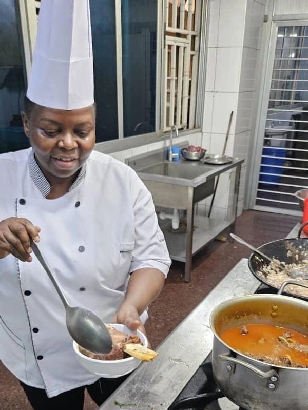 Chef serving food in a kitchen wearing a white uniform and hat.