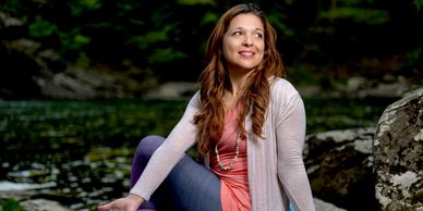 A woman in casual clothes sits by a river, smiling and looking away.