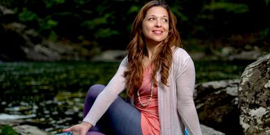A woman in casual clothes sits by a river, smiling and looking away.