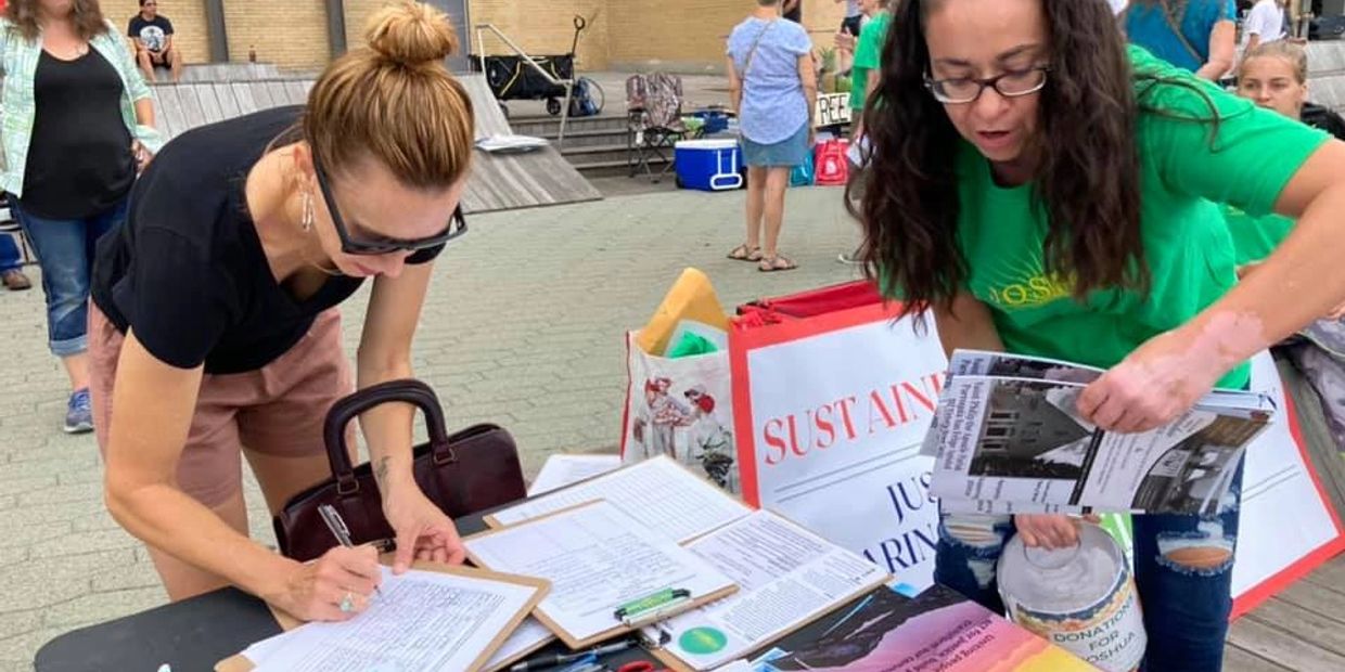 Two women stand near a table with papers. One is writing something and another is holding papers.