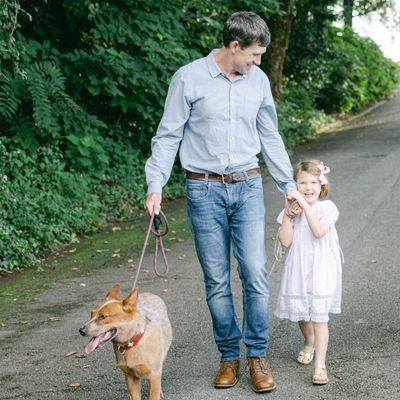 Father and daughter walking their dog on a tree-lined path.