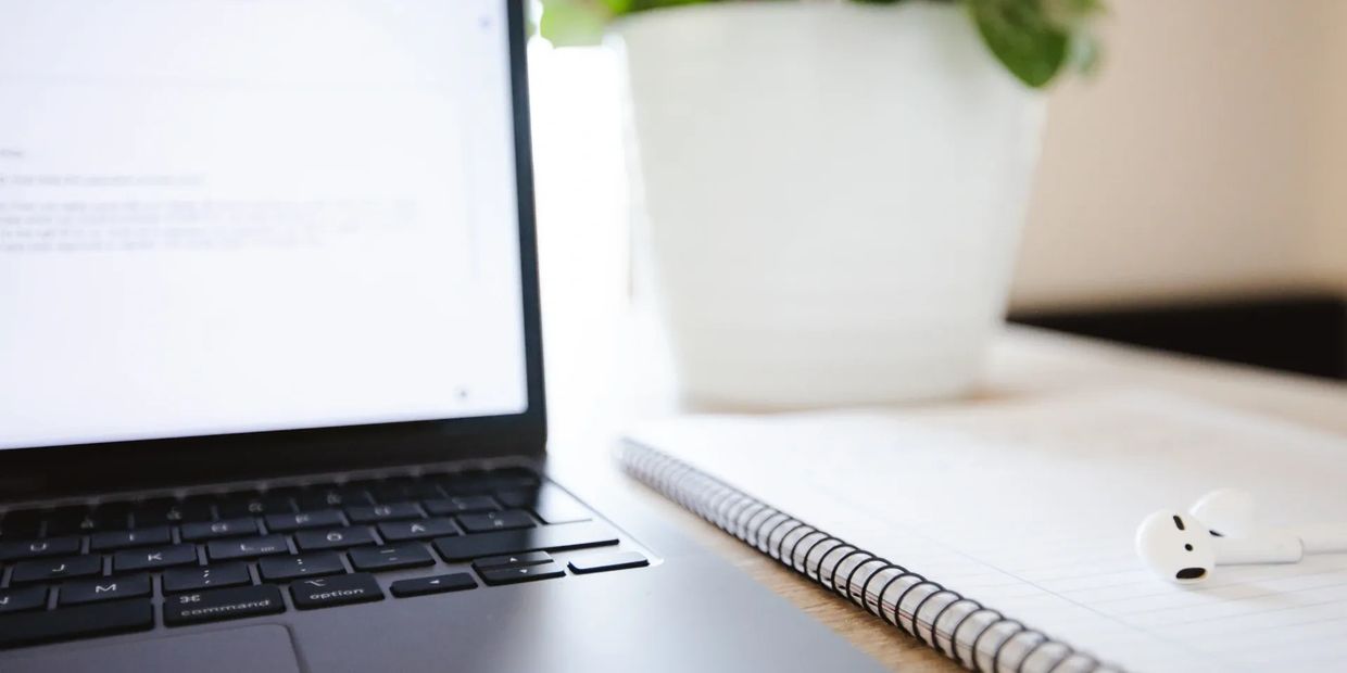 A laptop on a desk with a notebook and ear buds