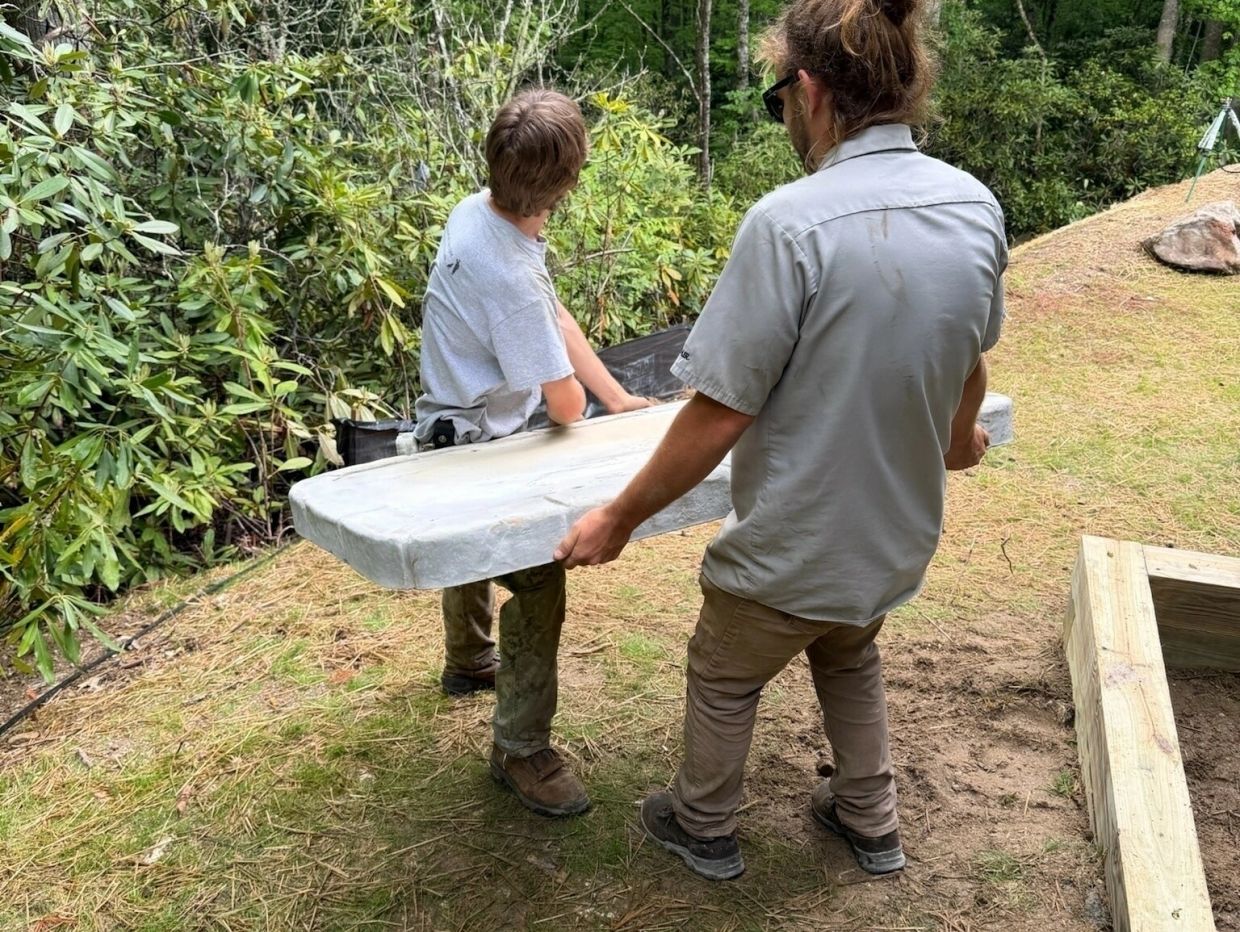 Two men carrying a heavy object outdoors in a wooded area.