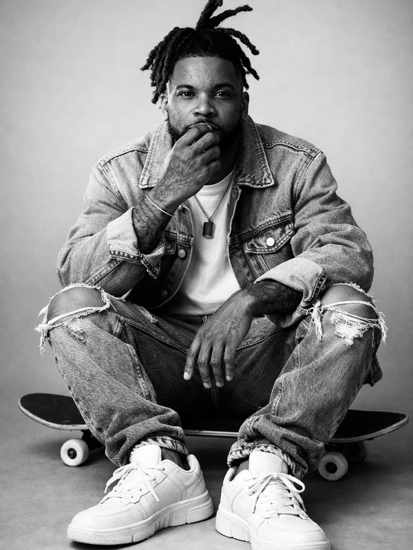 Man in denim jacket and ripped jeans sitting on a skateboard.