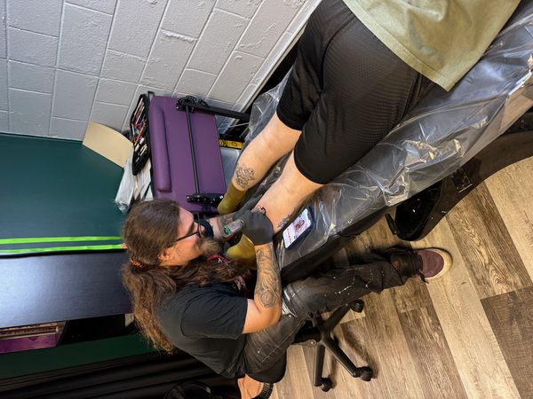 Tattoo artist working on a customer's leg tattoo in a studio.