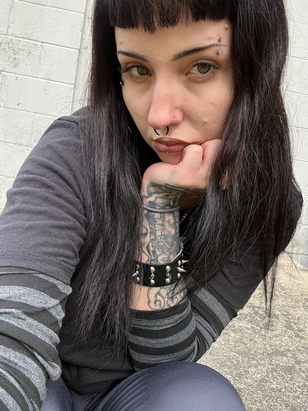 Person with black hair, piercings, and tattoos posing thoughtfully outdoors.