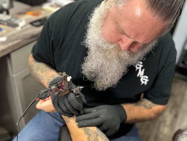 A tattoo artist with a long beard working on a client's arm.
