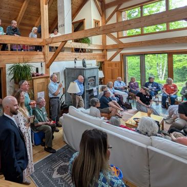 A large group gathered in a spacious, sunlit living room with wooden beams and large windows.