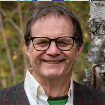 Smiling man with glasses outdoors, wearing a sweater and collared shirt.