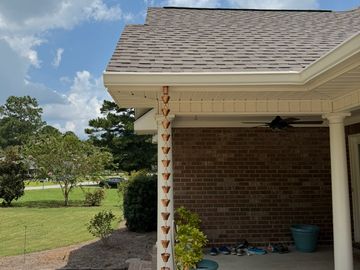 Rain chain hanging from porch roof on a sunny day with green lawn and blue sky.