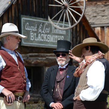 Three men in vintage western attire converse outside a blacksmith shop.