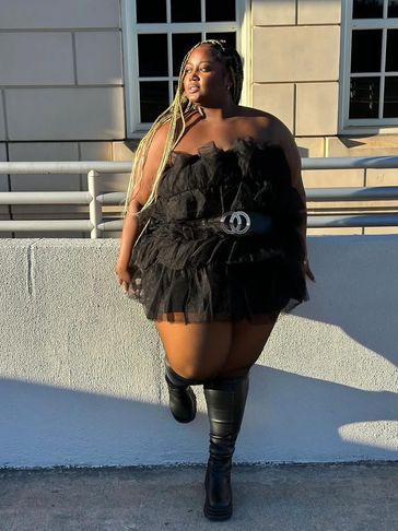 Confident woman in a black ruffled dress and knee-high boots poses outdoors.