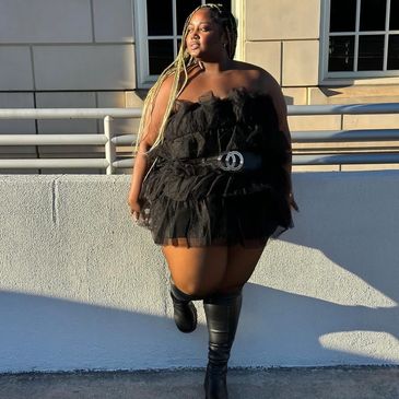 Confident woman in a black ruffled dress and knee-high boots poses outdoors.