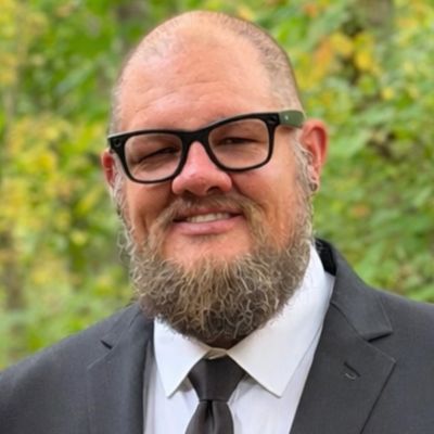 Smiling man with a beard and black glasses wearing a black suit 