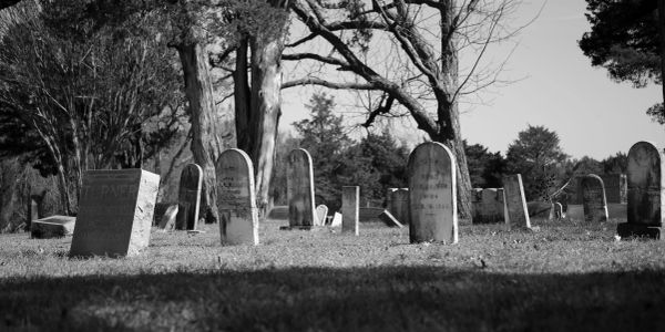 Black and white image of old gravestones in a cemetery with leafless trees.