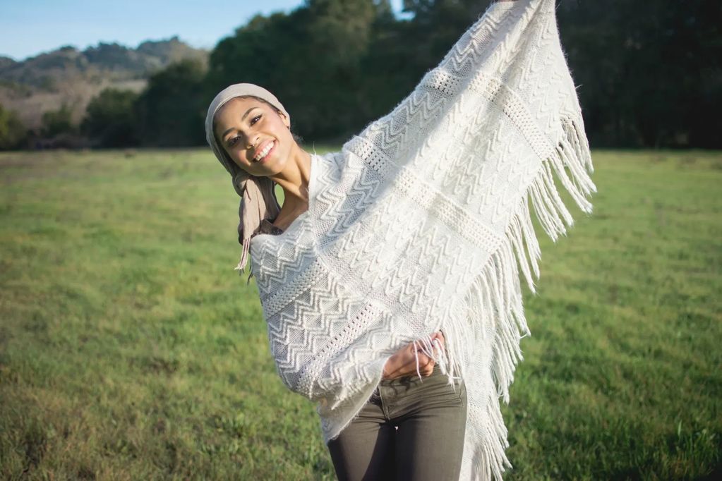A smiling woman outdoors holding a white fringed shawl.
