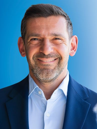 Smiling man in a blue suit jacket and white shirt against a blue background.