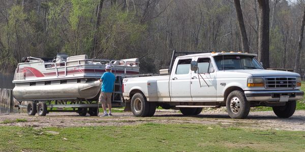 White pickup truck towing a pontoon boat on a trailer with a man nearby.