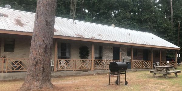 A rustic cabin porch with a grill and picnic tables in a wooded area.