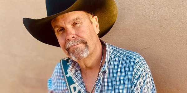 Man in a cowboy hat and checkered shirt posing against a beige wall.