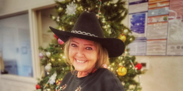 A smiling woman wearing a black cowboy hat in front of a decorated Christmas tree.