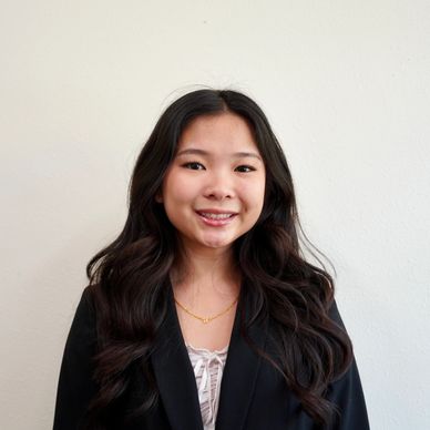 Young woman with long dark hair wearing a black blazer and gold necklace, smiling at the camera.