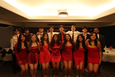Group of young adults dressed formally, women in red dresses and men in shirts with ties, posing indoors.