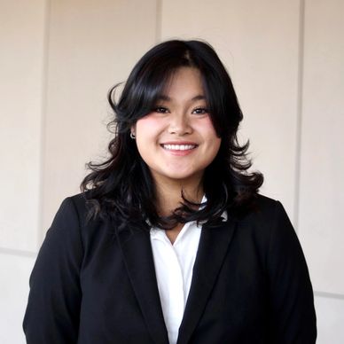 A young woman with black hair, wearing a black blazer and white shirt, smiling confidently.