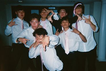 A group of seven young men in white shirts and pink ties posing with hand gestures indoors.