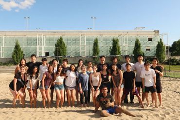 Large group of young people posing on a sandy volleyball court on a sunny day.