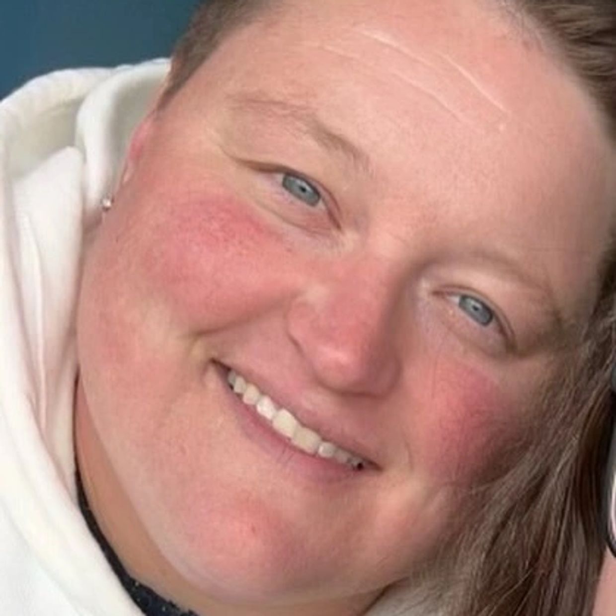 Close-up of a smiling person with blue eyes and light skin wearing a white hoodie.