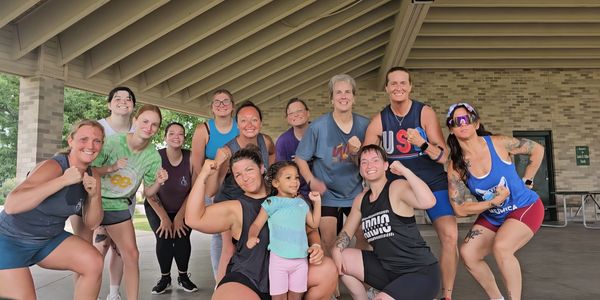 Group of women and a child flexing muscles together in a covered outdoor area.