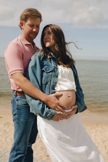 A couple embraces on the beach, highlighting the woman's pregnant belly.