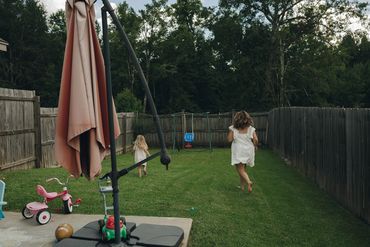 Two children running in a fenced backyard with toys and an umbrella stand.