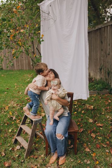 Mother holds baby while toddler kisses her on cheek outdoors.