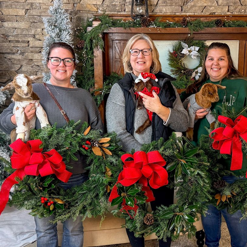Three women holding Christmas wreaths and baby goats indoors.