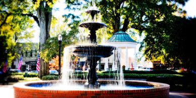 Ornate water fountain in a sunny park surrounded by trees and flags.