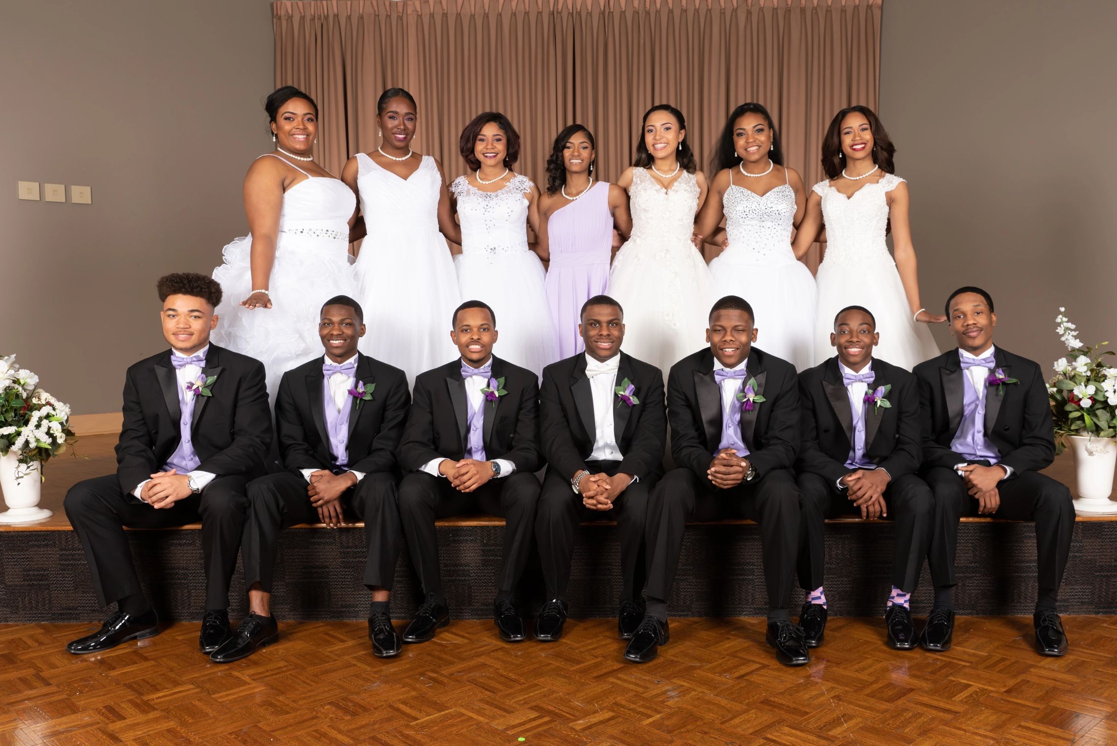 Group portrait of young men in tuxedos and women in white dresses at a formal event.