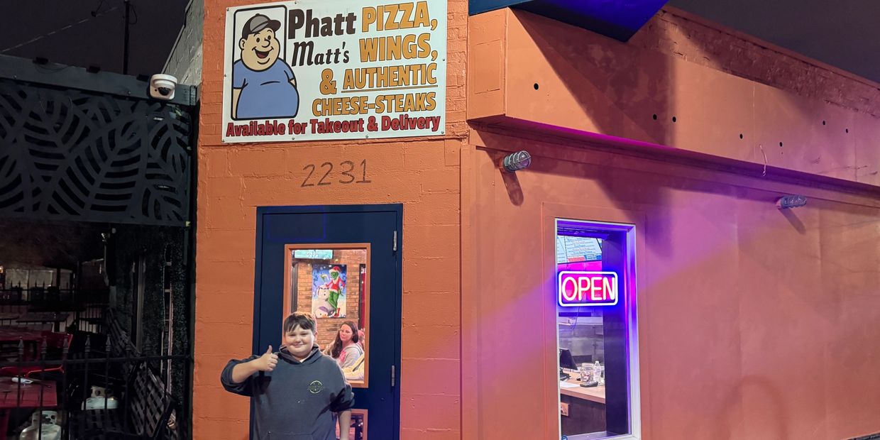 A boy giving a thumbs-up outside Phatt Matt's pizza place at night.