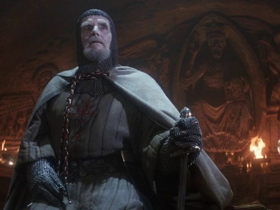 A medieval knight in chainmail and cloak holds a sword in a dark, torch-lit chamber.