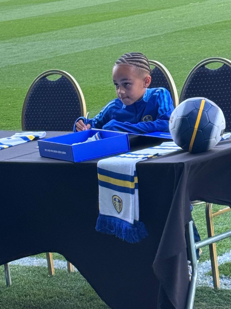 A young boy in a blue sports jacket signing an autograph at a table with a soccer ball and scarf.