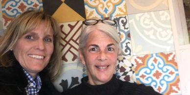 Two women smiling in front of a colorful tiled wall.