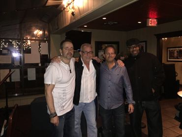 Four men posing together inside a dimly lit room with warm lighting.