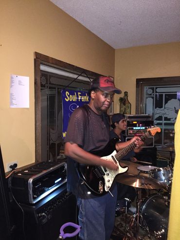 Musician playing electric guitar indoors with drummer in background.